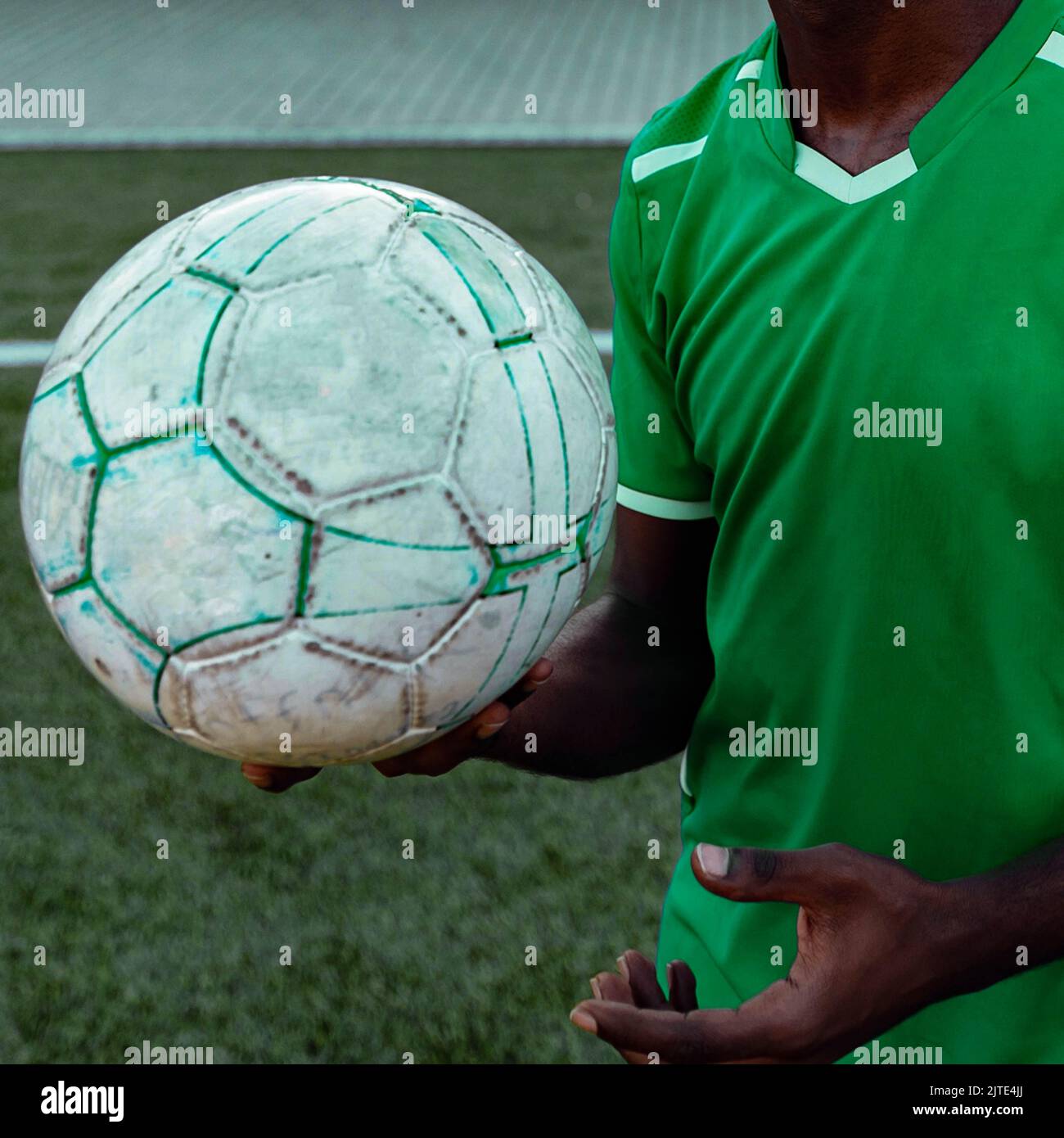 Primo piano di un uomo dai colori scuri e irriconoscibili in un'uniforme da calcio verde che tiene una palla da calcio su un campo da calcio Foto Stock