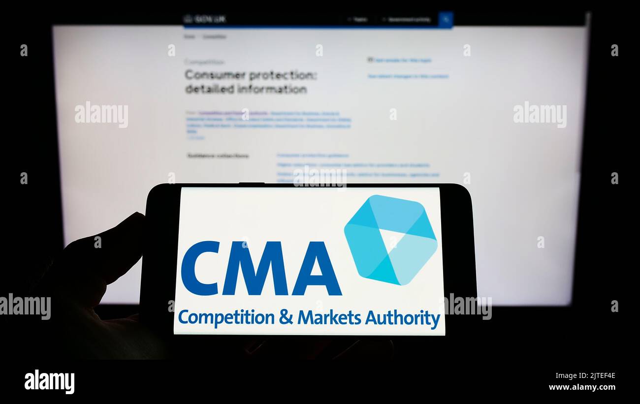 Persona che tiene il cellulare con il logo della British Competition and Markets Authority (CMA) sullo schermo di fronte alla pagina web. Messa a fuoco sul display del telefono. Foto Stock