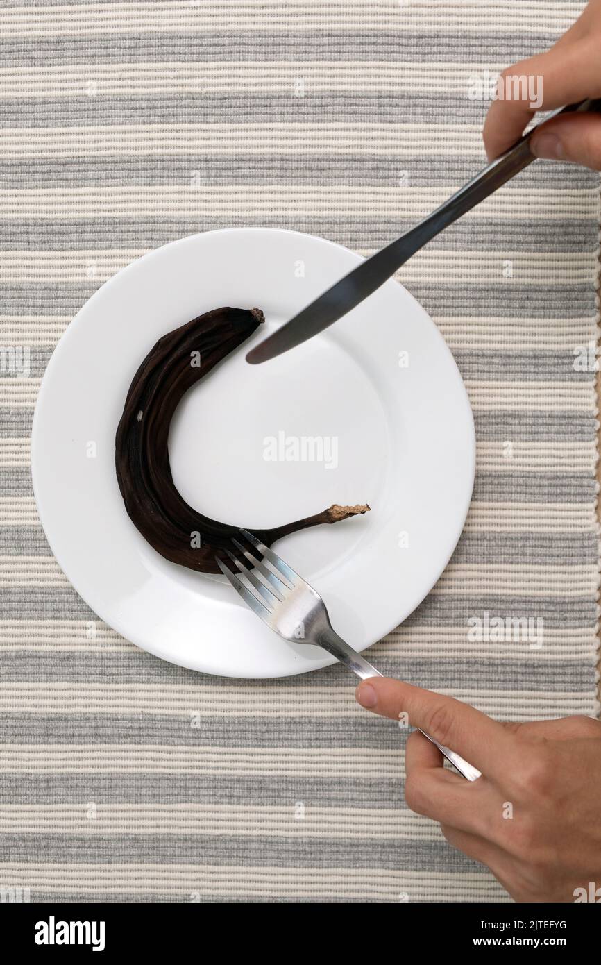 L'uomo con forchetta e coltello si prepara a mangiare banana nera ammuffita secca su piatto bianco. Non abbastanza cibo nel tempo affamato. Foto Stock