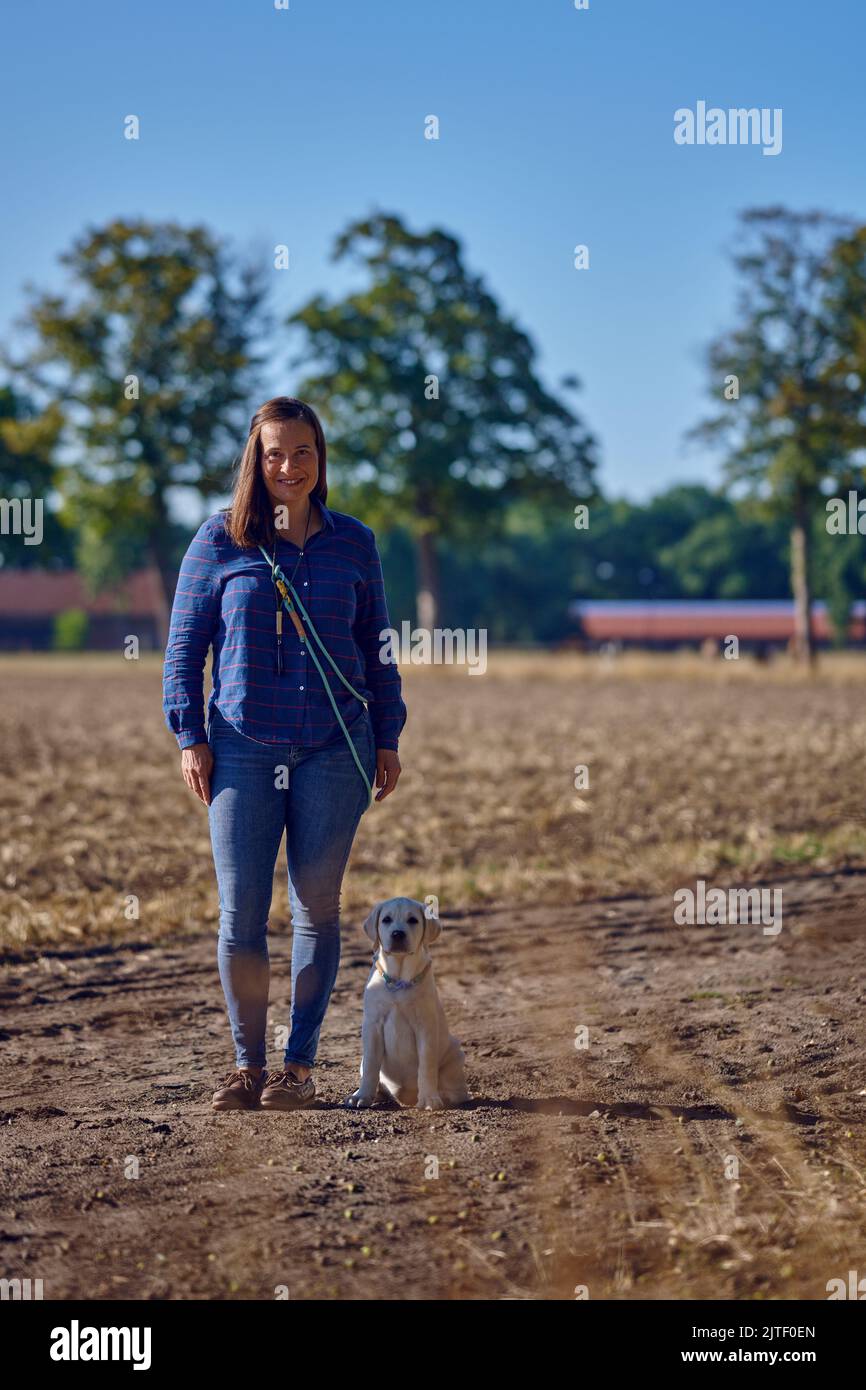 Bruna felice donna di mezza età in piedi con il suo piccolo cucciolo Labrador mentre starnutisce su un sentiero rurale Foto Stock