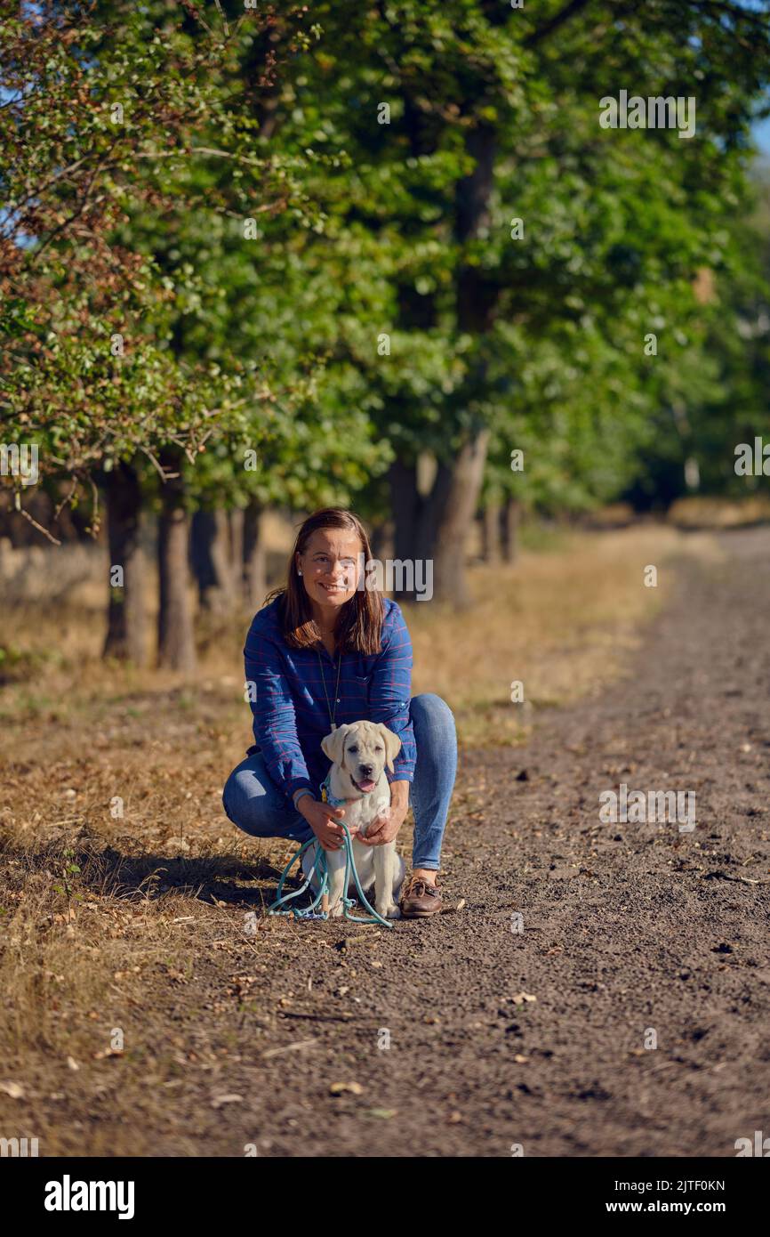 Felice brunetta donna di mezza età che starnutisce con il suo piccolo cucciolo Labrador mentre starnutisce su un sentiero rurale Foto Stock