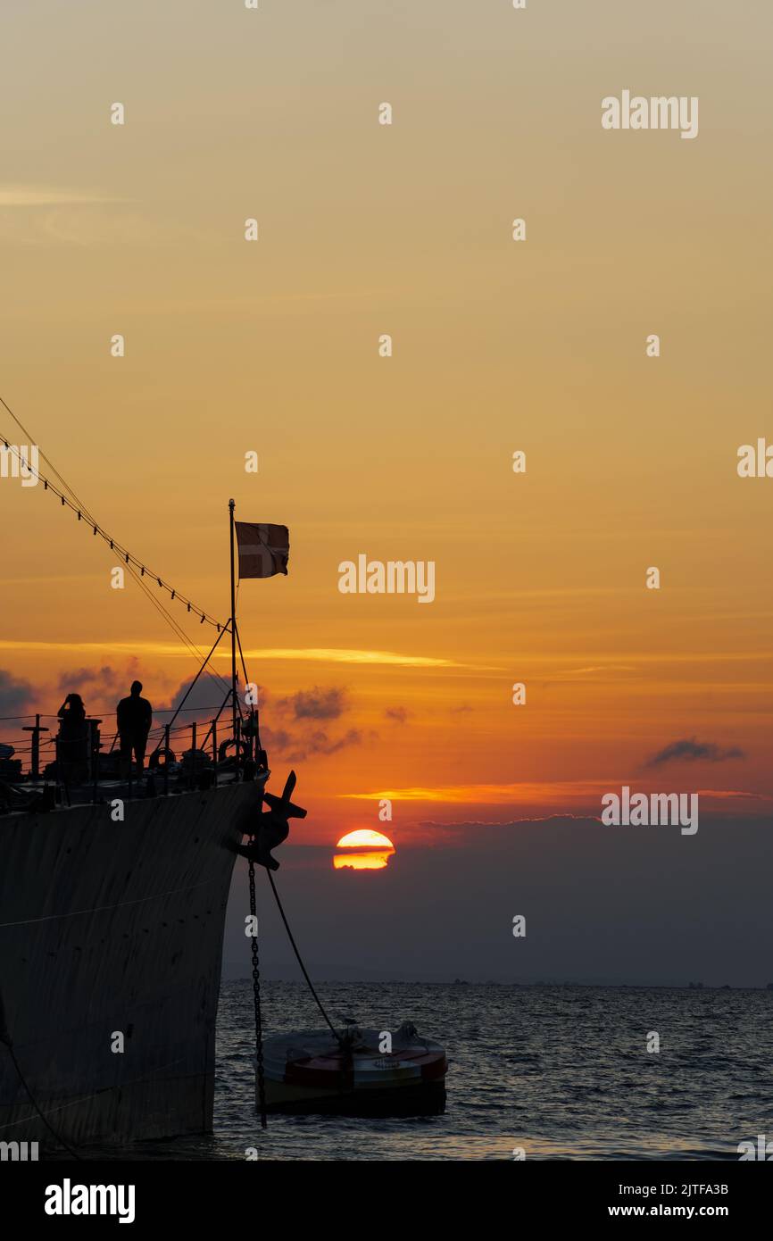 Vista serale della vecchia nave da guerra greca ormeggiata con bandiera ondulata e silhouette umane a bordo, contro il tramonto sul livello del mare. Foto Stock