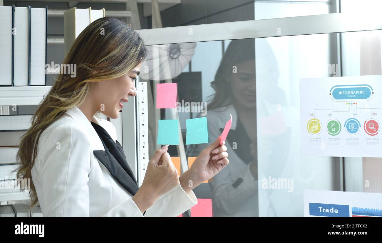 Felice donna asiatica busimess guardando la carta stick su vetro con momento di successo in ufficio Foto Stock
