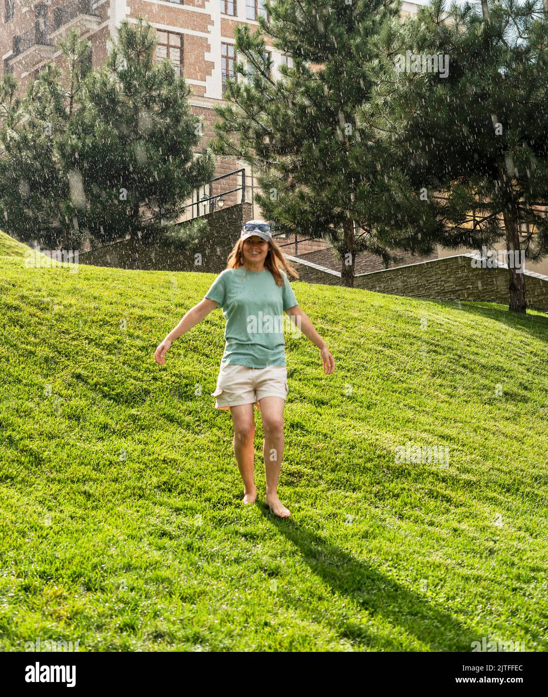 Felice giovane donna a piedi nudi camminare sul prato verde prato godendo la pioggia calda in estate godendo la sensazione di natura Foto Stock