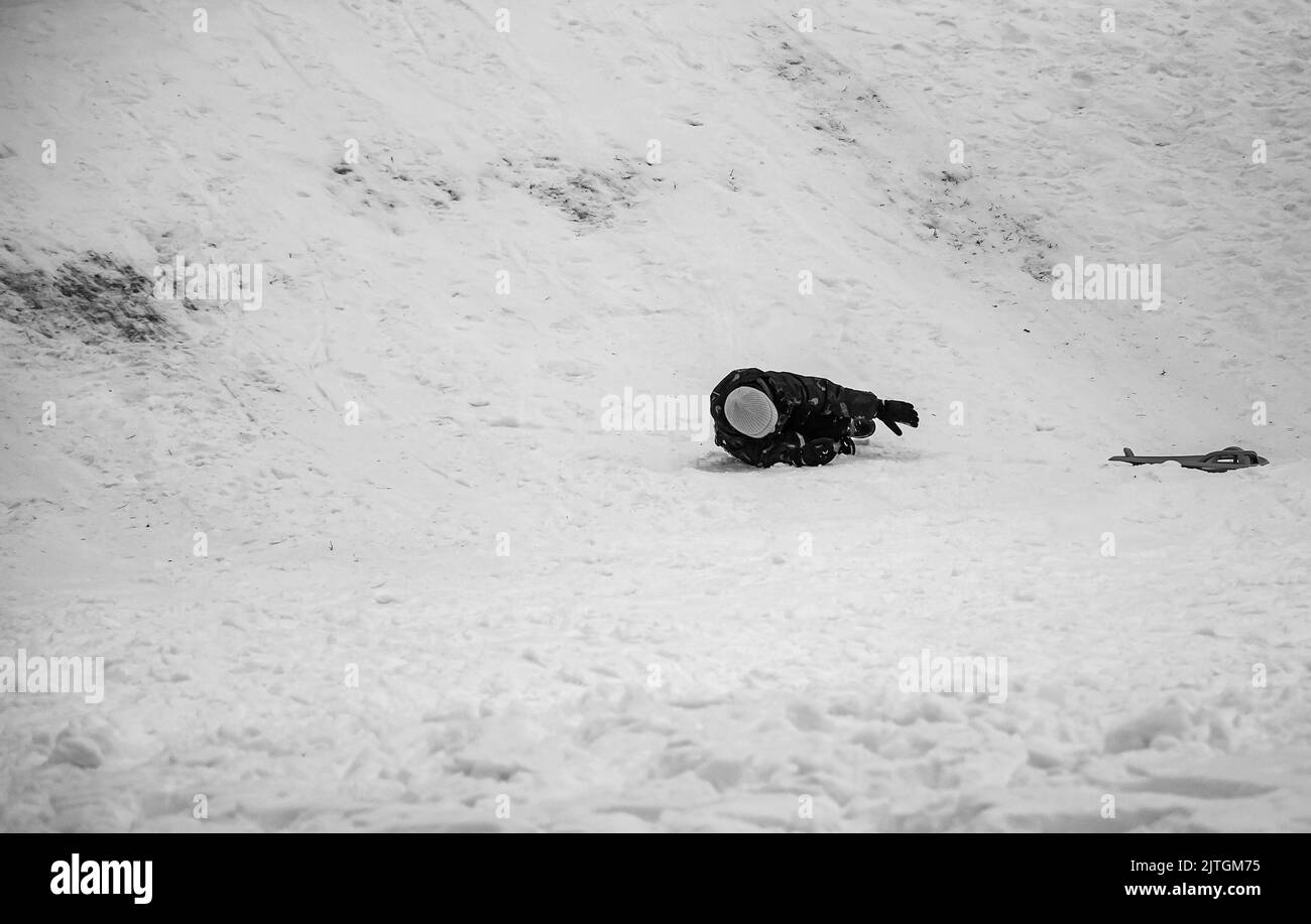 Il ragazzino slittava in discesa in inverno. Il ragazzo cade nella neve. Giochi all'aperto per bambini. Slitte per bambini in un parco coperto di neve. Infanzia, Foto Stock