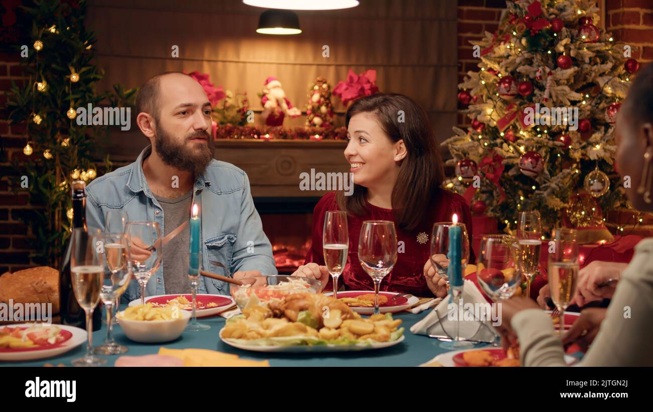 Coppia festosa che festeggia il Natale al tavolo della cena con parenti stretti. Persone positive che si siedono nel soggiorno per celebrare le autentiche vacanze invernali tradizionali con i propri cari. Foto Stock