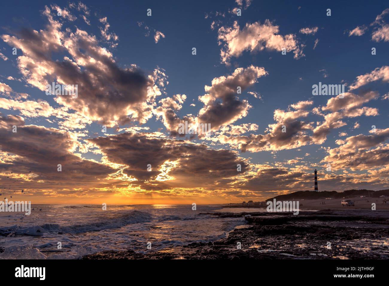 Spettacolare cielo al tramonto con nuvole e riflessi sul mare. Un faro bianco e nero sulla destra. Foto Stock