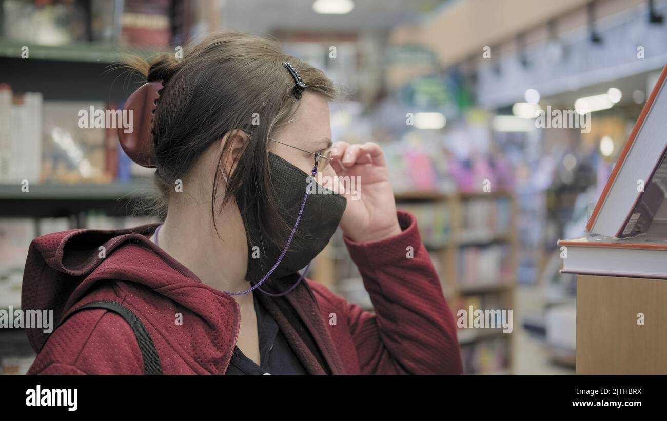 Donna che indossa occhiali e maschera contro il virus sceglie libro. Lady regola gli occhiali Foto Stock