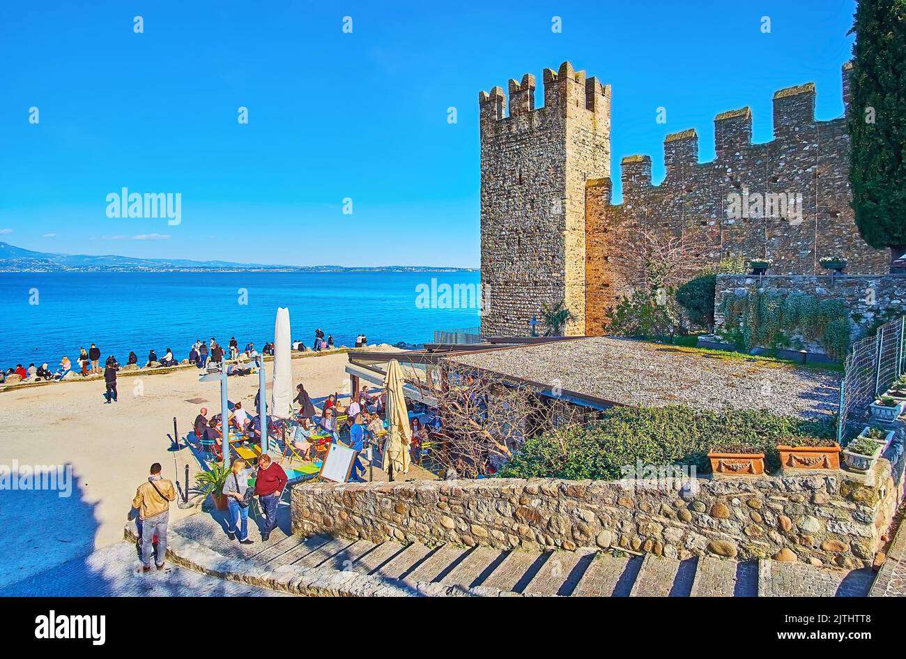 SIRMIONE, ITALIA - 10 APRILE 2022: Le mura medievali e la torre del Castello Scaligero con la spiaggia della Spiaggia del Prete e le acque blu del Lago di Garda, in Apri Foto Stock