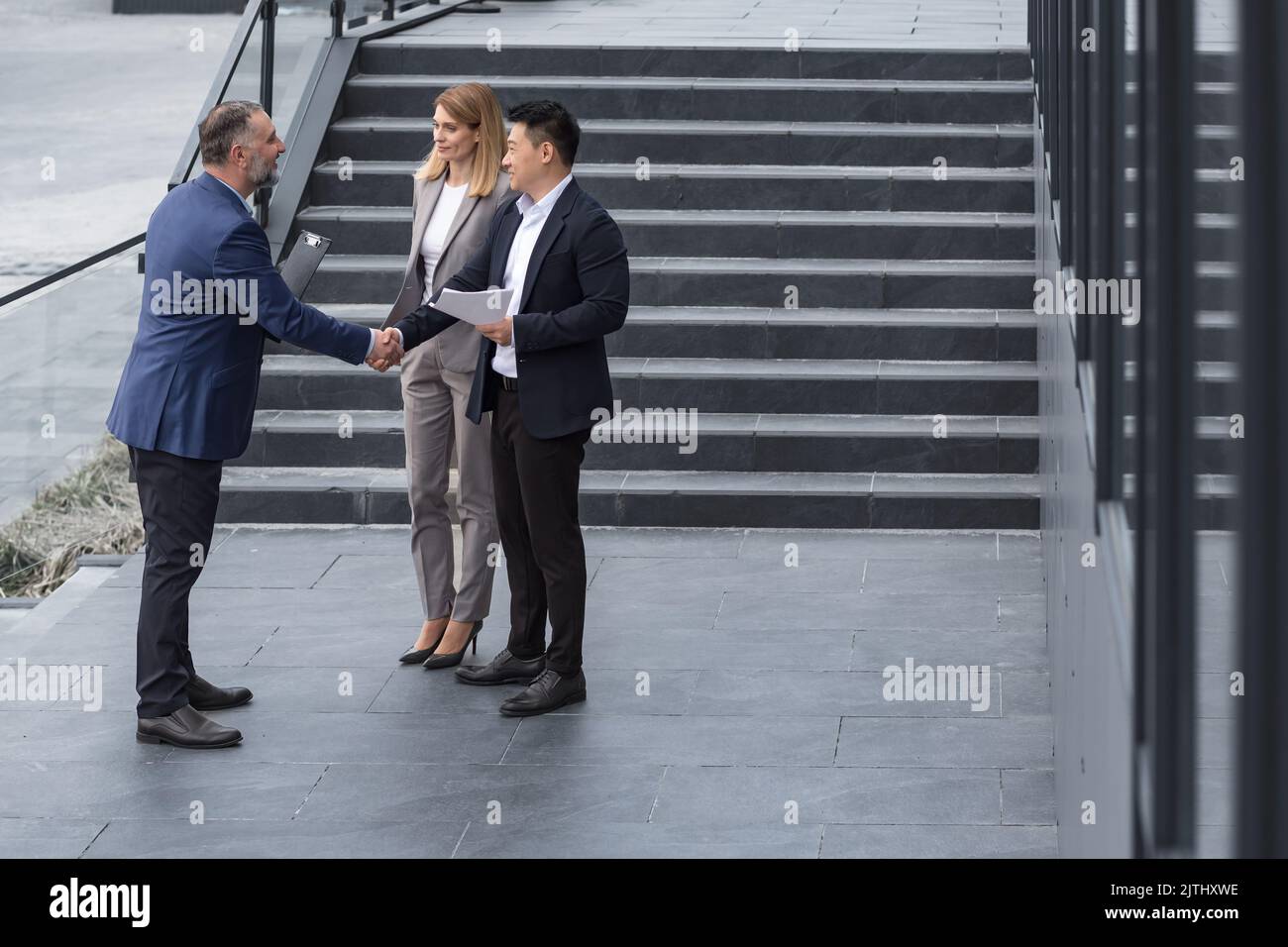 Incontro di tre colleghi esterni all'edificio dell'ufficio, specialisti IT esperti e maturi, accoglienza e scuotimento delle mani, persone aziendali in tute aziendali, gruppi diversi di persone Foto Stock
