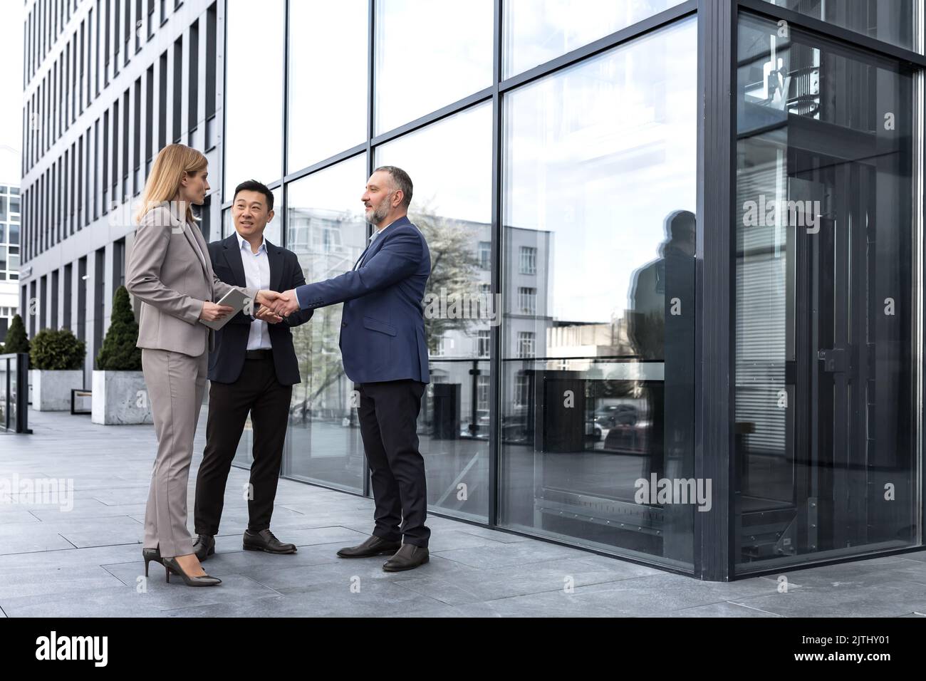 Incontro di tre colleghi esterni all'edificio dell'ufficio, specialisti IT esperti e maturi, accoglienza e scuotimento delle mani, persone aziendali in tute aziendali, gruppi diversi di persone Foto Stock