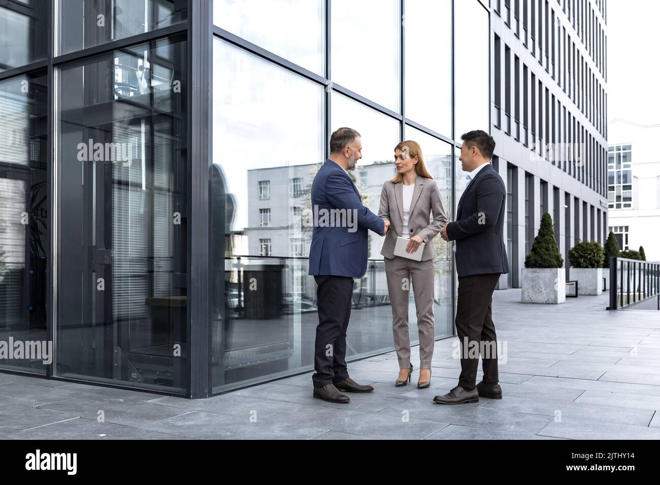 Incontro di tre colleghi esterni all'edificio dell'ufficio, specialisti IT esperti e maturi, accoglienza e scuotimento delle mani, persone aziendali in tute aziendali, gruppi diversi di persone Foto Stock