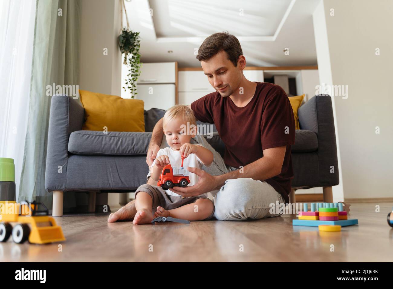 Papà e figlio premurosi giocano con il giocattolo dell'auto seduto sul pavimento a casa, godendosi il tempo trascorso insieme. Legame di famiglia. Padre giorno. Infanzia spensierata. Felice paternità. Sviluppo del bambino Foto Stock