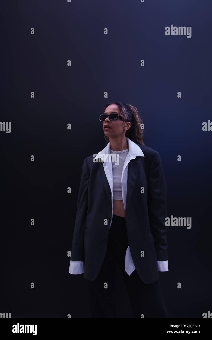 elegante donna afroamericana in occhiali da sole e tuta oversize su blu scuro Foto Stock