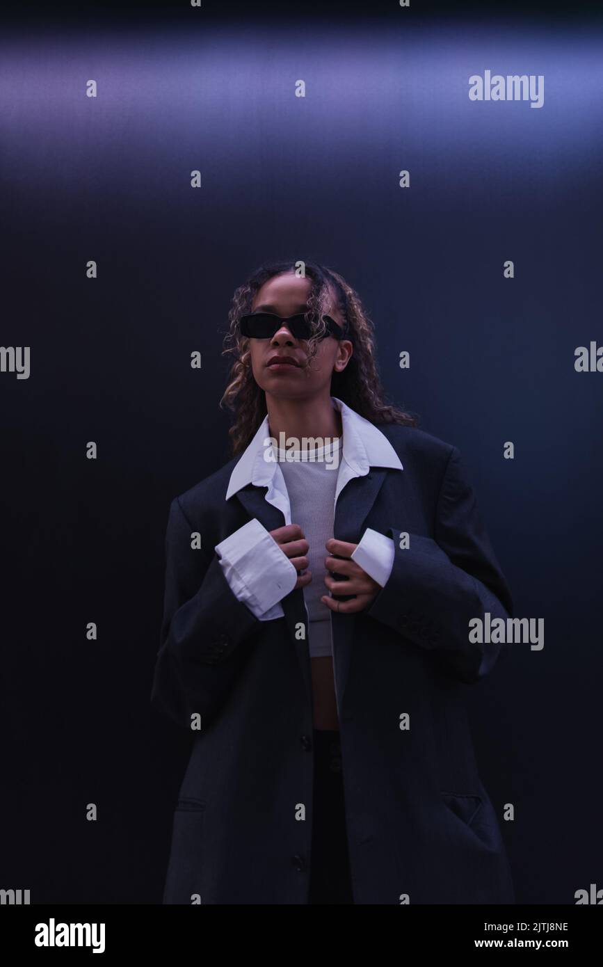 curly afroamericana donna in occhiali da sole regolazione blazer oversize vicino a parete blu scuro Foto Stock