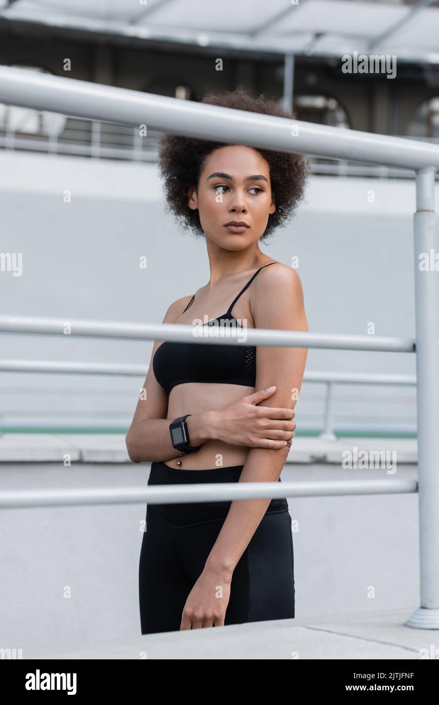 donna afro-americana snella in abbigliamento sportivo nero e fitness tracker guardando lontano vicino recinzione Foto Stock