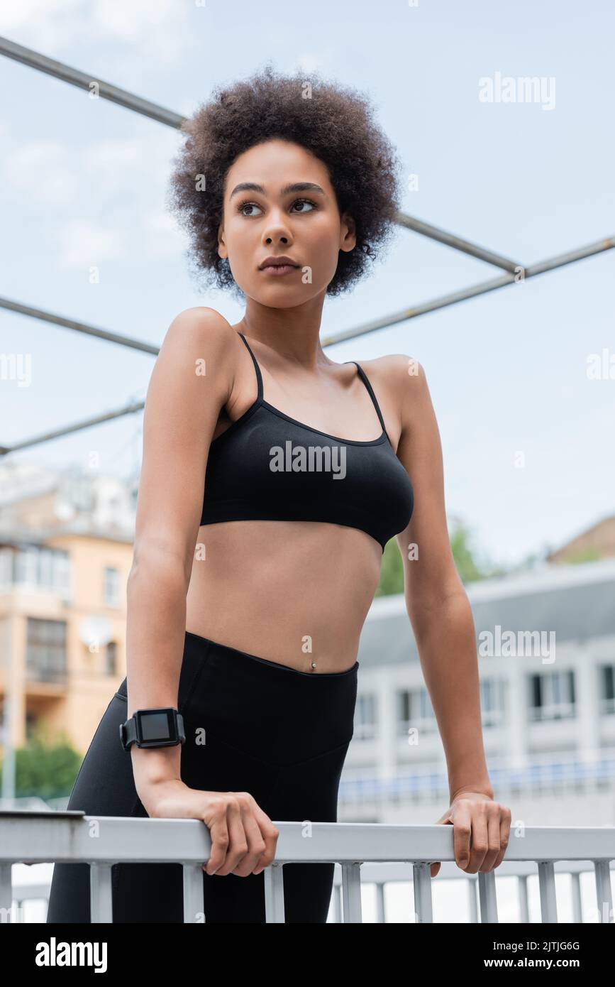 curly afroamericana donna in bra nera sport e fitness tracker guardando lontano vicino recinzione Foto Stock