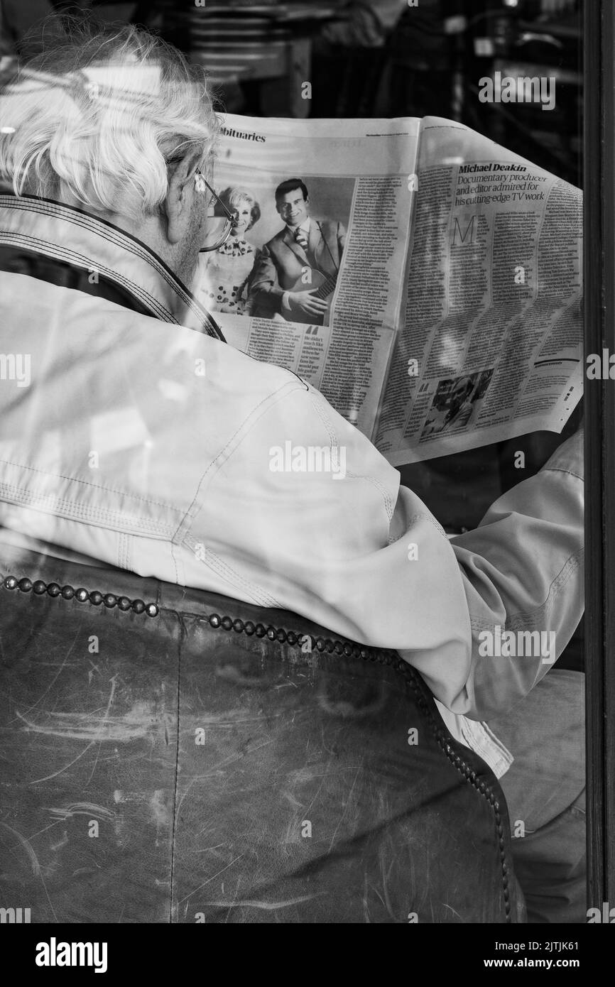 Fotografia di strada di un vecchio che legge un giornale in un caffè, presa attraverso la finestra del caffè Foto Stock