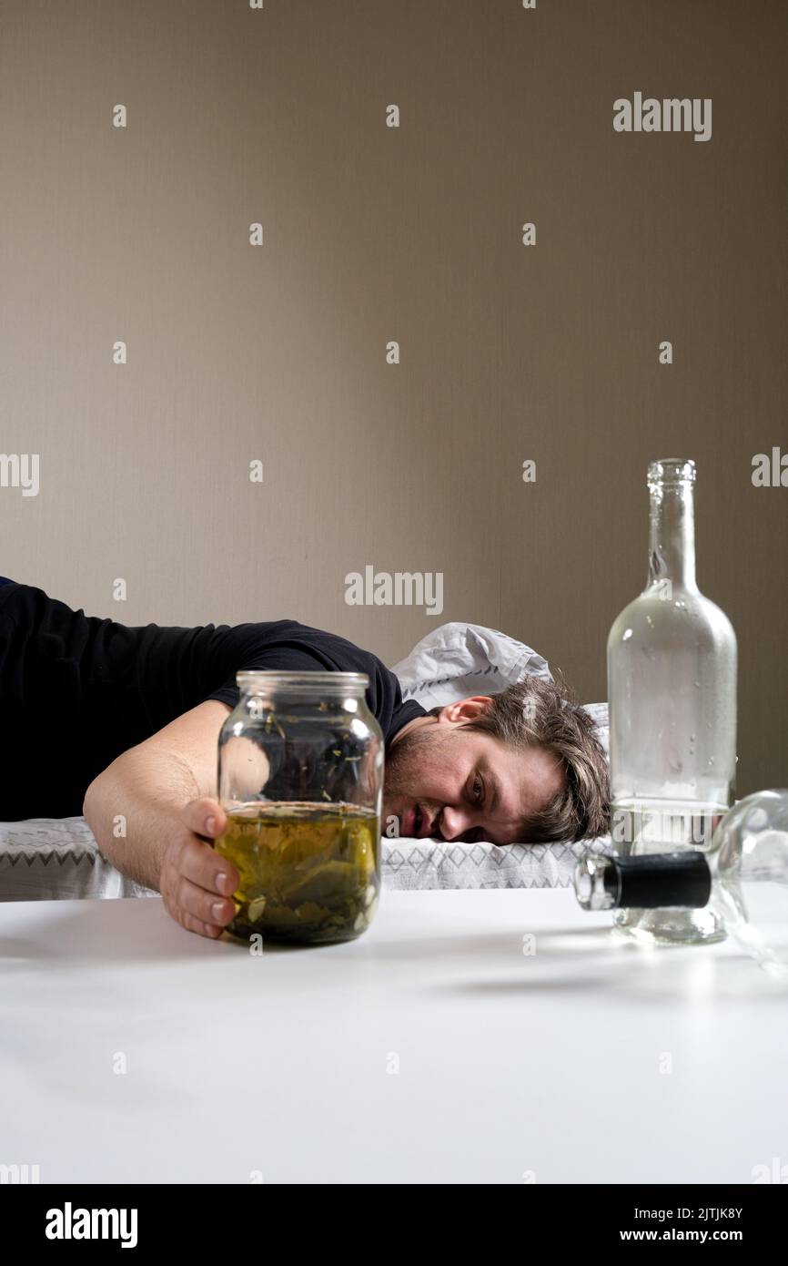 Un uomo con una dipendenza da alcol si trova sul letto e raggiunge con la mano un vaso di cetriolo salato salamoia. Rimedi popolari per i hangovers Foto Stock