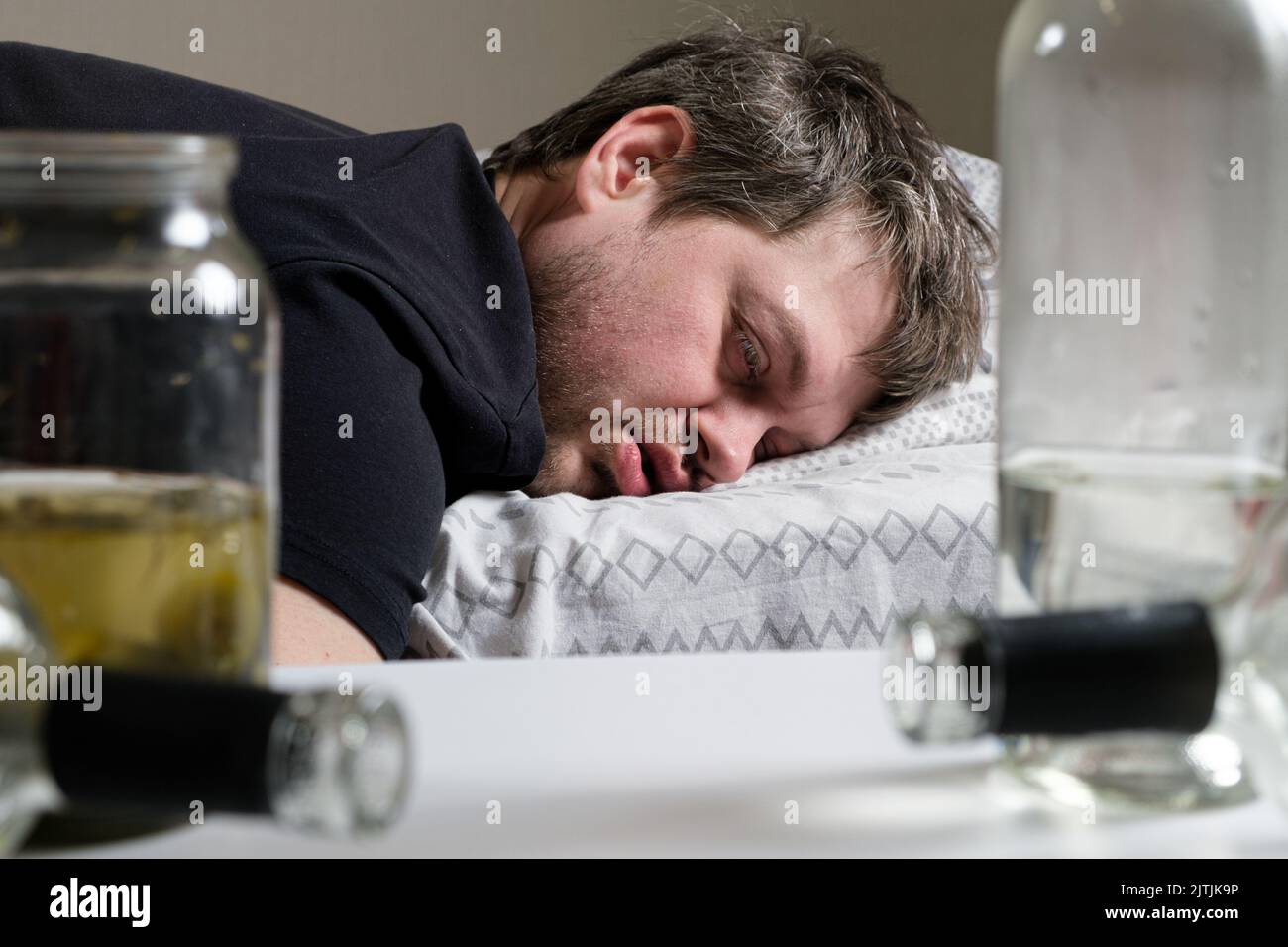 Un alcolista ubriaco si trova sul letto, bottiglie vuote sul tavolo. Alcolismo, dipendenza da alcol come problema sociale Foto Stock