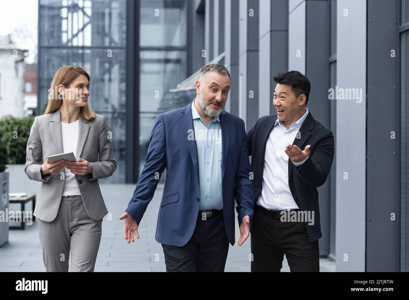 Un team diversificato di specialisti IT, responsabili di ingegneri senior ed esperti, un gruppo di tre lavoratori che si girano felicemente fuori da un edificio di uffici, colleghi in tute aziendali. Foto Stock