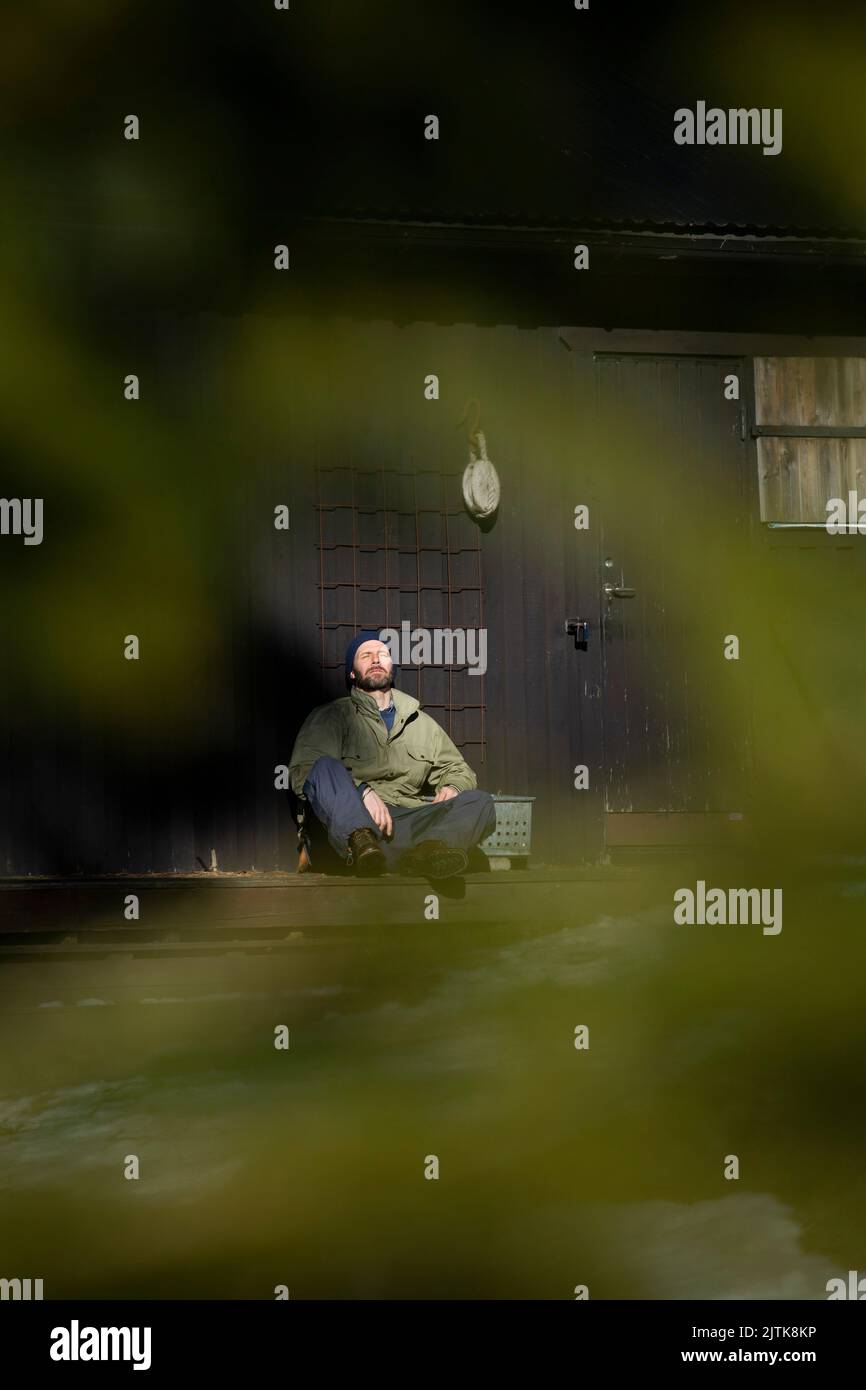 Intera lunghezza di uomo maturo seduto con gli occhi chiusi sul portico durante il giorno di sole Foto Stock