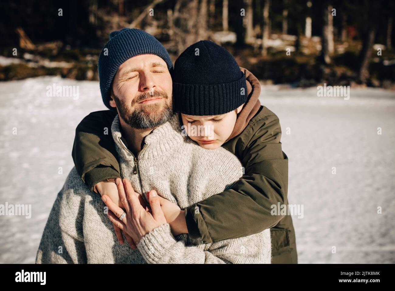 Ragazzo in abiti caldi che abbraccia il padre con gli occhi chiusi durante la giornata di sole in inverno Foto Stock