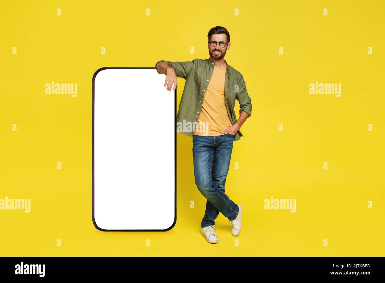 Corpo intero dell'uomo appoggiato sul grande smartphone con schermo bianco vuoto, in piedi su sfondo giallo studio, mockup Foto Stock
