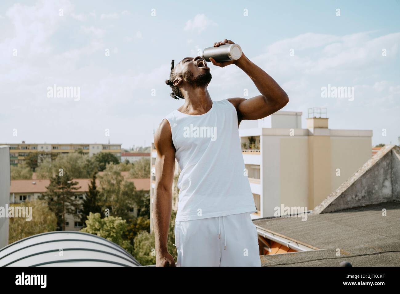 Giovane uomo che beve acqua dalla bottiglia mentre si trova in piedi sul tetto durante le giornate di sole Foto Stock