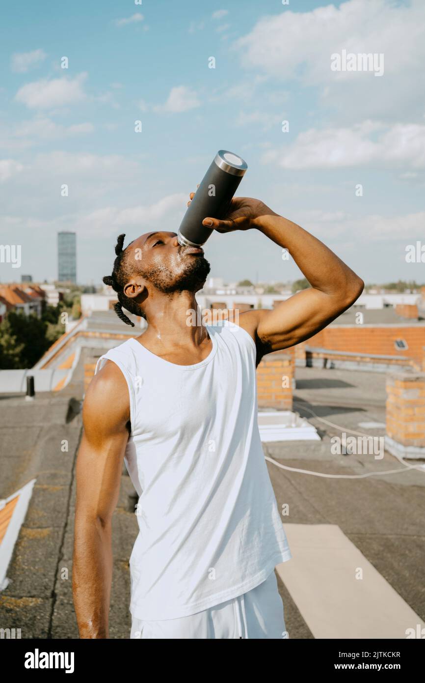 Giovane uomo che beve acqua dalla bottiglia sul tetto durante la giornata di sole Foto Stock