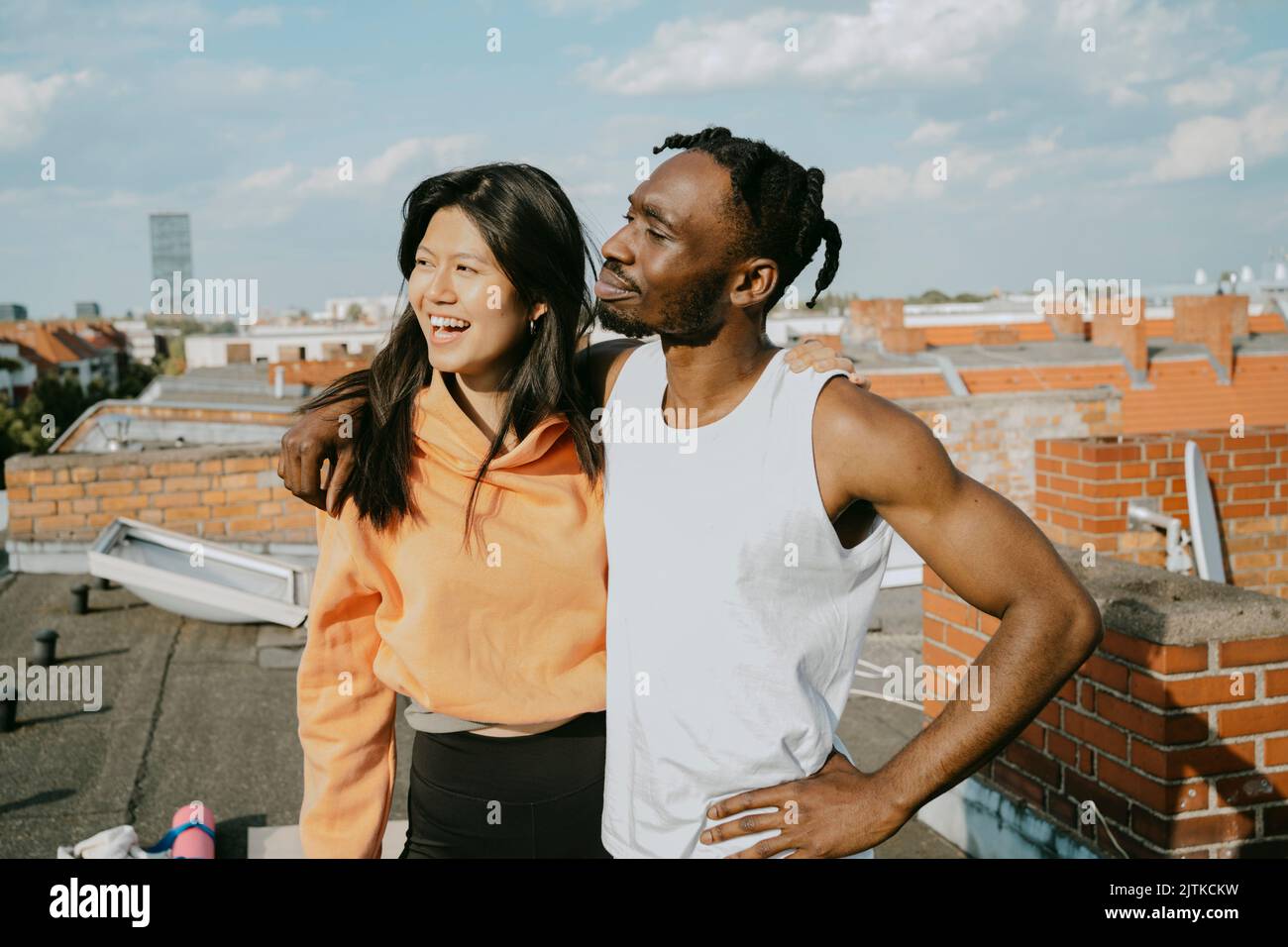 Giovane uomo sorridente in piedi con un braccio attorno a un'amica che guarda lontano mentre si trova sul tetto Foto Stock