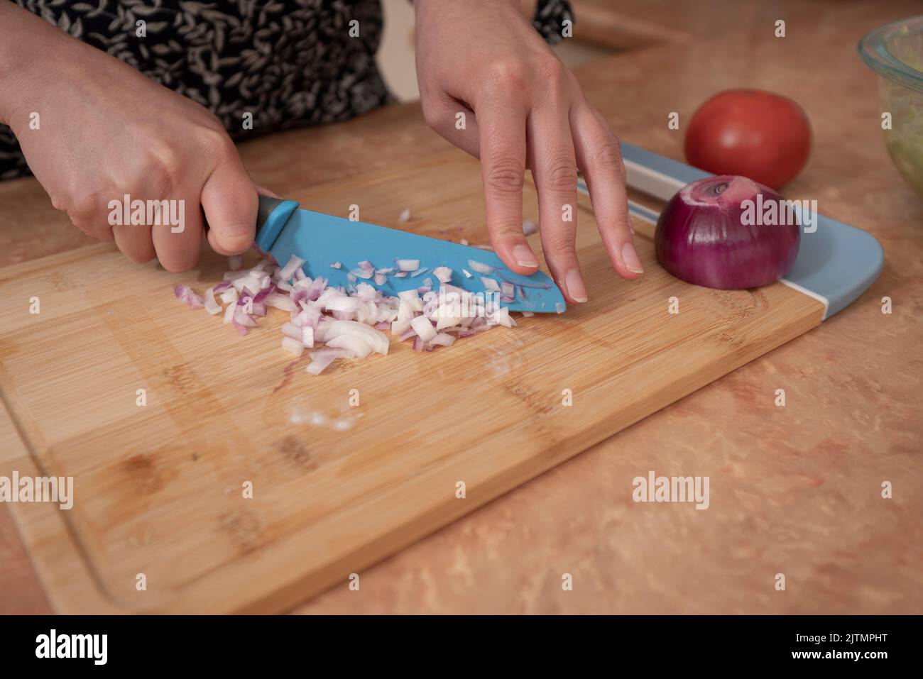 Primo piano delle mani di una donna che trita le cipolle con un coltello blu su un asse di legno al bancone della cucina Foto Stock