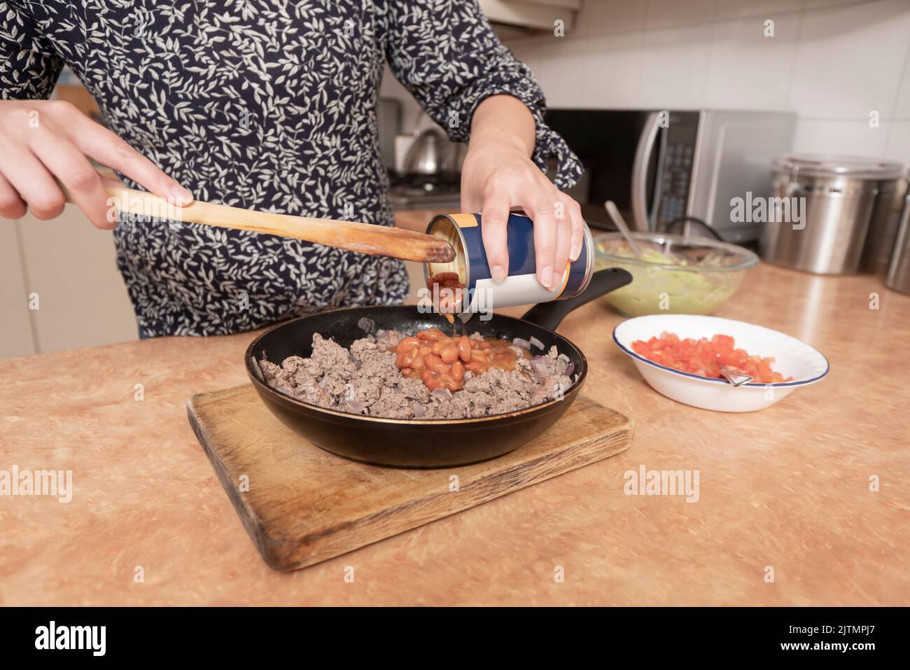 Primo piano delle mani della donna svuotare una lattina di fagioli con carne macinata utilizzando un cucchiaio di legno in una padella di metallo nero Foto Stock