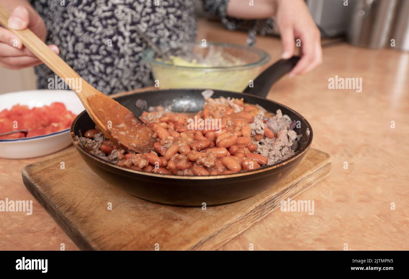 Primo piano delle mani di una donna che mescola fagioli con carne macinata usando un cucchiaio di legno in una padella di metallo nero Foto Stock