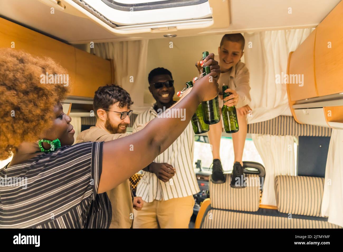Gruppo interrazziale di allegri amici che brindano con bottiglie di birra, bevendo un drink per celebrare un'occasione speciale all'interno del loro comodo e spazioso camper van. Trascorrere insieme i fine settimana in un campeggio estivo in pulmino. Foto di alta qualità Foto Stock
