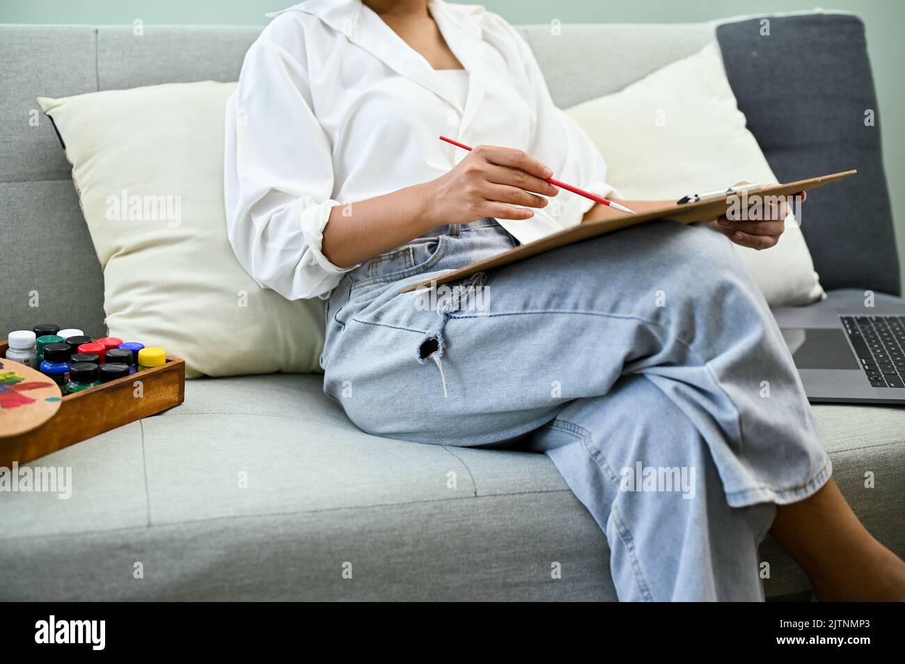 Una donna asiatica rilassata si siede sul divano nel suo salotto di casa, disegnando e dipingendo il colore dell'acqua, rendendo la sua bella opera d'arte. Concetto creativo di hobby Foto Stock