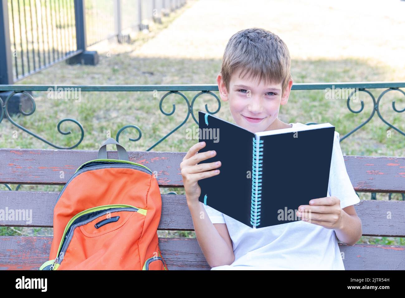 Un ragazzo adorabile che legge un libro, un notebook seduto su una panca per strada. Prepararsi per una lezione a scuola. Concetto di ritorno a scuola. Foto Stock