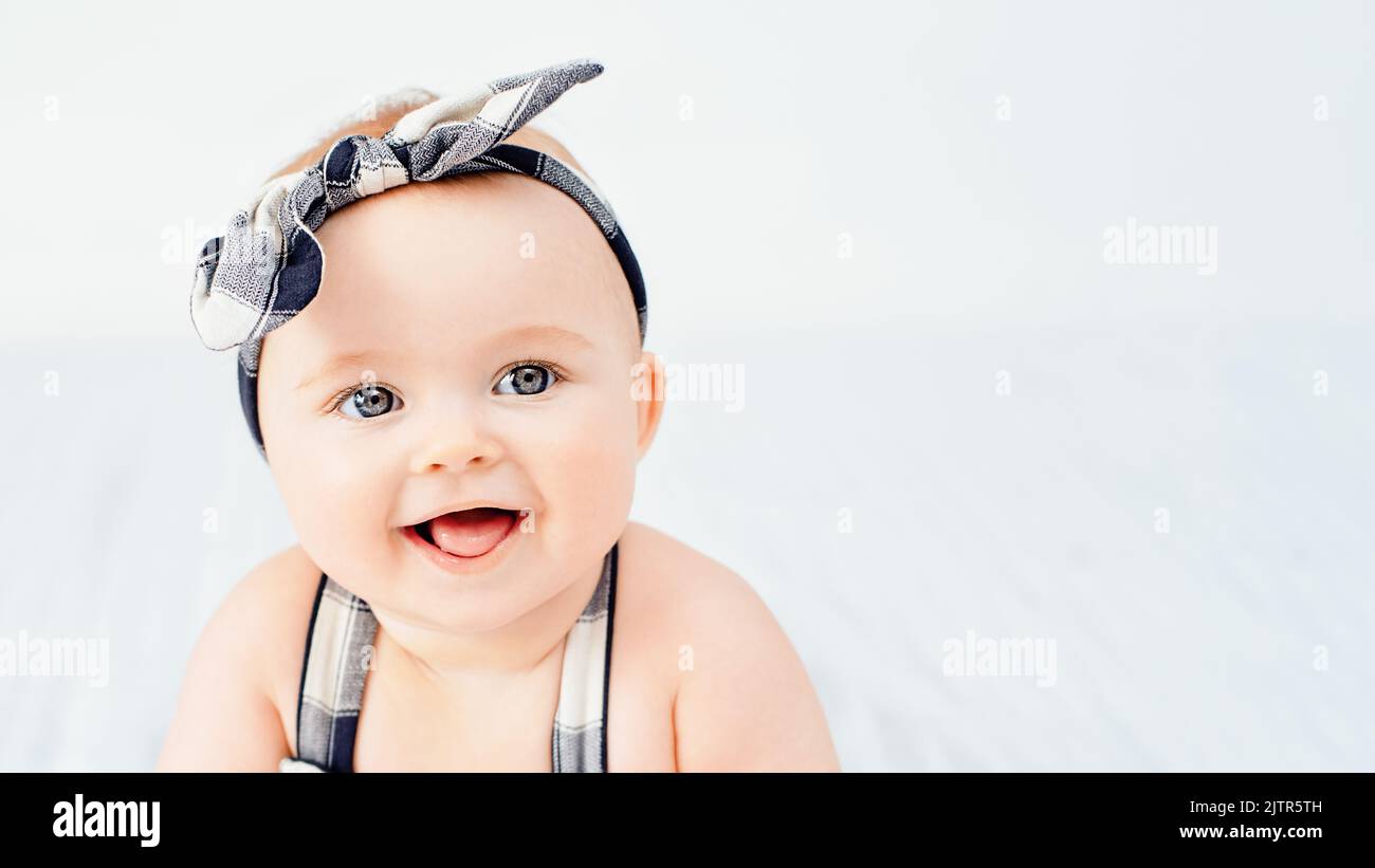 Bambino di sette mesi seduto sul letto. Bambina sorridente carina su coperta morbida bianca. Ragazza che indossa la fascia. Incantevole bambino dagli occhi blu. Spazio di copia. Foto Stock