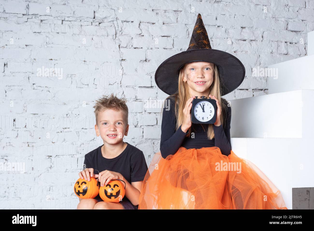 Halloween. I bambini in costumi e cappelli di strega creepy stanno tenendo una sveglia nera e un cestino a forma di zucca con le prelibatezze di Halloween mentre siedono Foto Stock