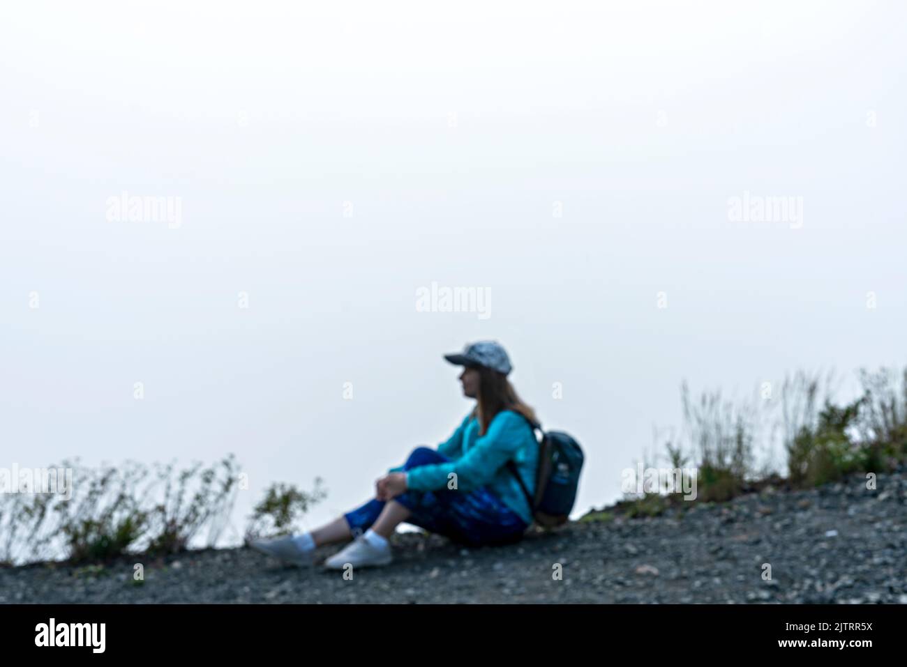 Giovane donna in berretto e vestiti sportivi blu con zaino seduto sul bordo della montagna contro le nuvole bianche e la nebbia guardando la vista turismo escursioni e a. Foto Stock