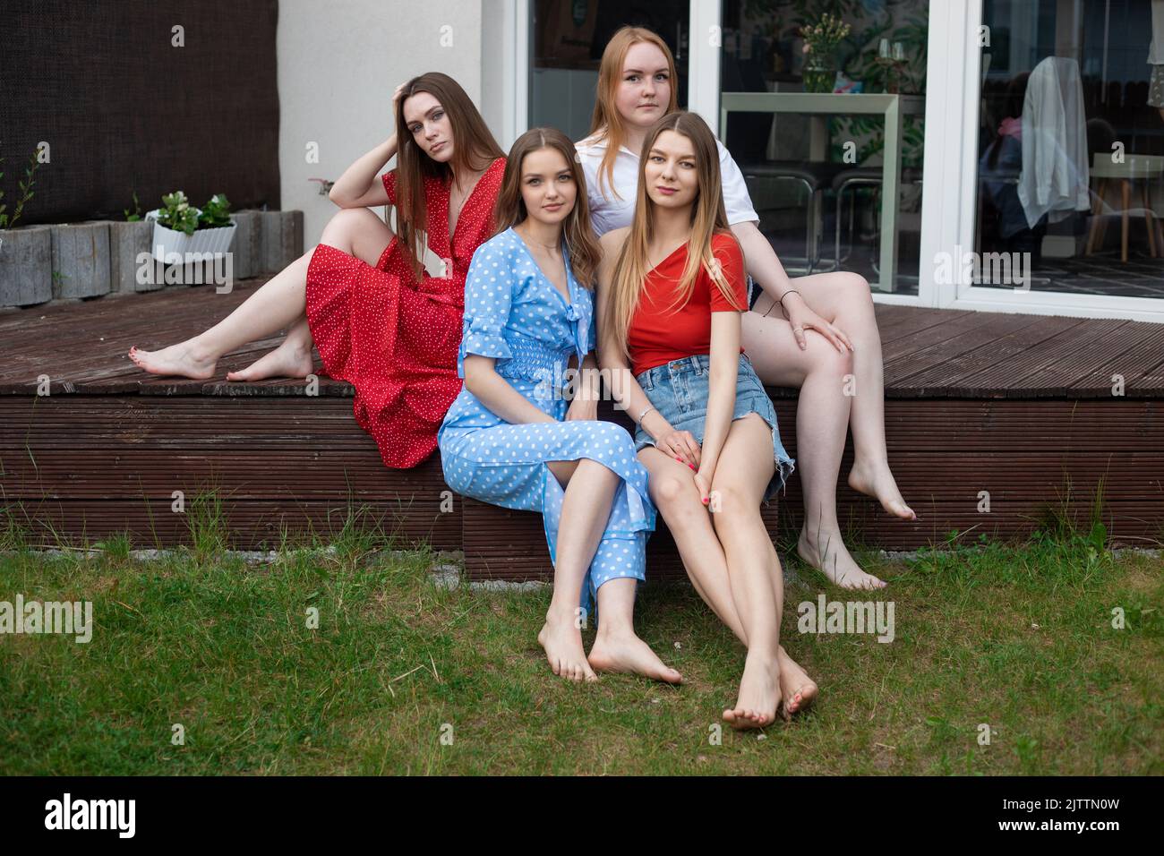 Gruppo di giovani donne a piedi nudi fiduciose e belle con lunghi capelli seduti su veranda di legno vicino all'erba verde, estate. Foto Stock