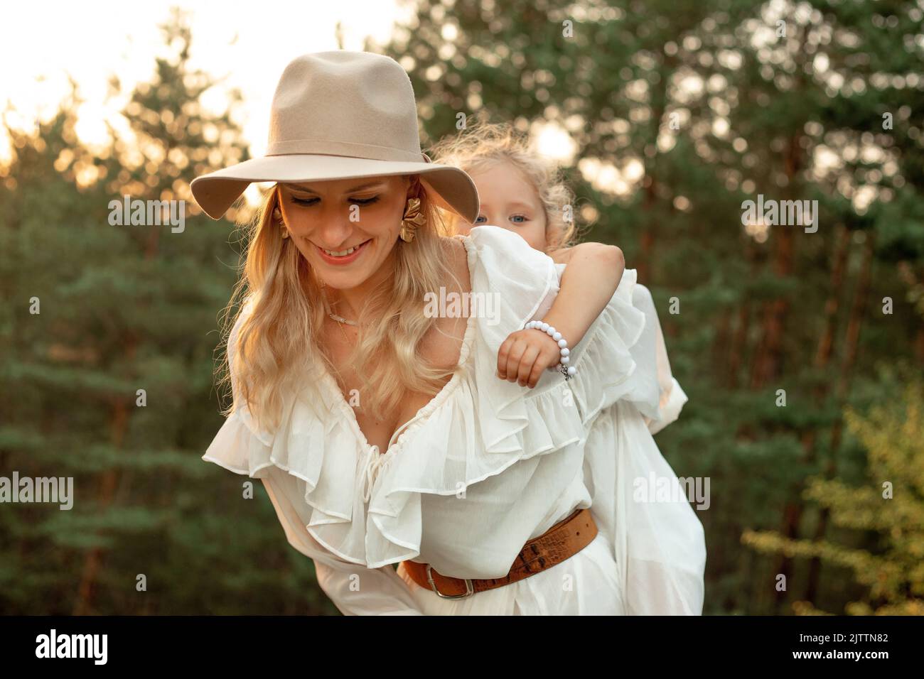 Ritratto di donna felice adorabile madre che dà cavalcata piggyback a bambina in foresta illuminata dal tramonto in estate. Foto Stock