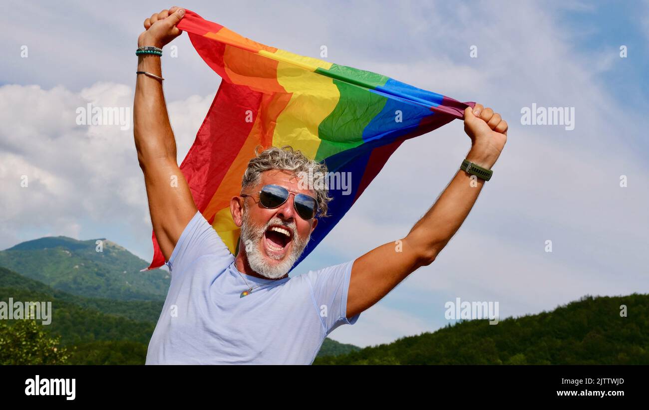 Ritratto di un anziano caucasico dai capelli grigi con una barba e occhiali da sole che reggono una bandiera arcobaleno LGBTQIA contro uno sfondo cielo, grida di protesta, celebra il mese di Pride in uscita Day Foto Stock