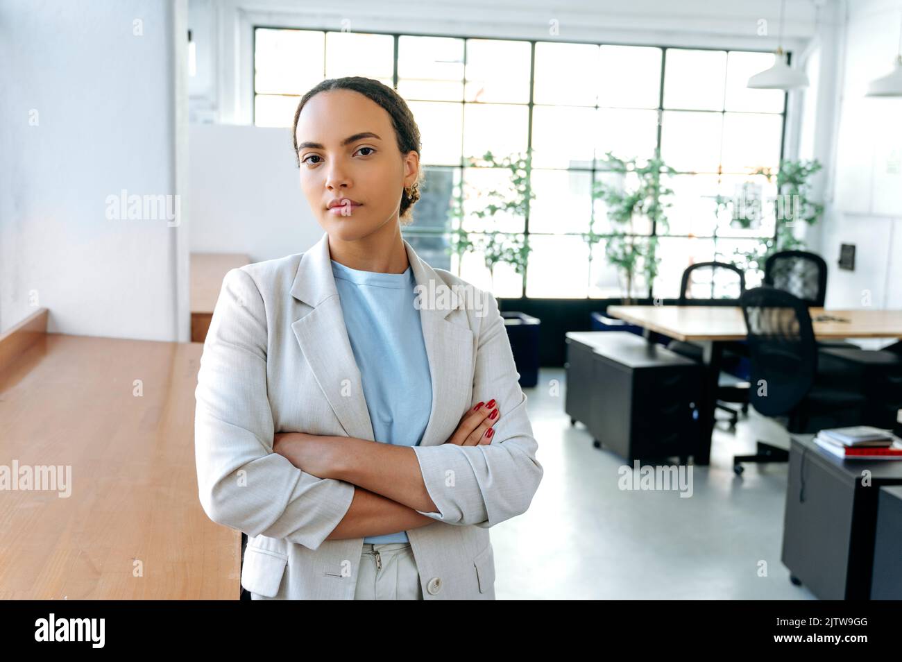Ritratto di grande successo influente giovane donna d'affari di razza mista, indossando abiti formali, in piedi in ufficio moderno con braccia incrociate, guardando la fotocamera Foto Stock