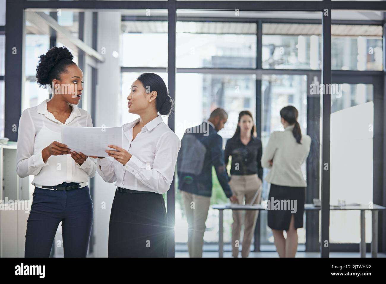 Mi fa molto piacere che ci abbia pensato: Due colleghi di lavoro femminili discutono di affari in ufficio mentre i loro colleghi lavorano in background. Foto Stock