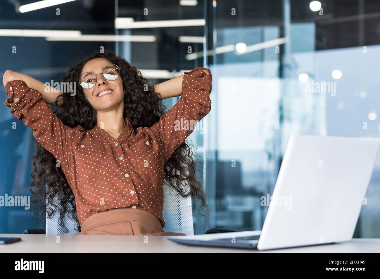 Giovane donna ingegnere sviluppatore programmatore lavora in ufficio all'interno della società IT, soddisfatto del risultato del lavoro, riposa ispanico guarda fuori dalla finestra, sorride le mani dietro la testa, sogni. Foto Stock