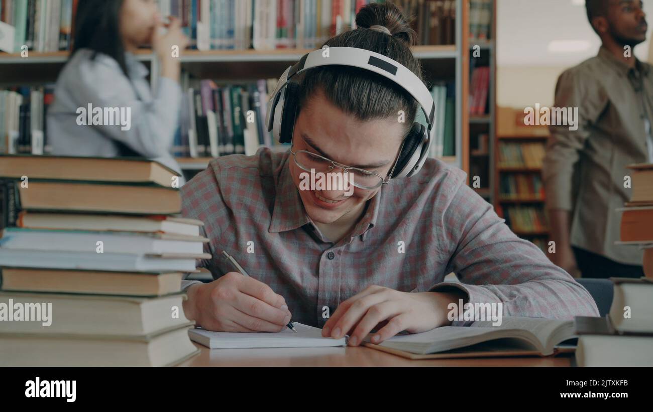 Giovane bel ragazzo caucasico seduto a tavola in grande biblioteca del college sta scrivendo qualcosa nel copybook. Sorride e sembra positivo e felice, libri sdraiati sul tavolo Foto Stock