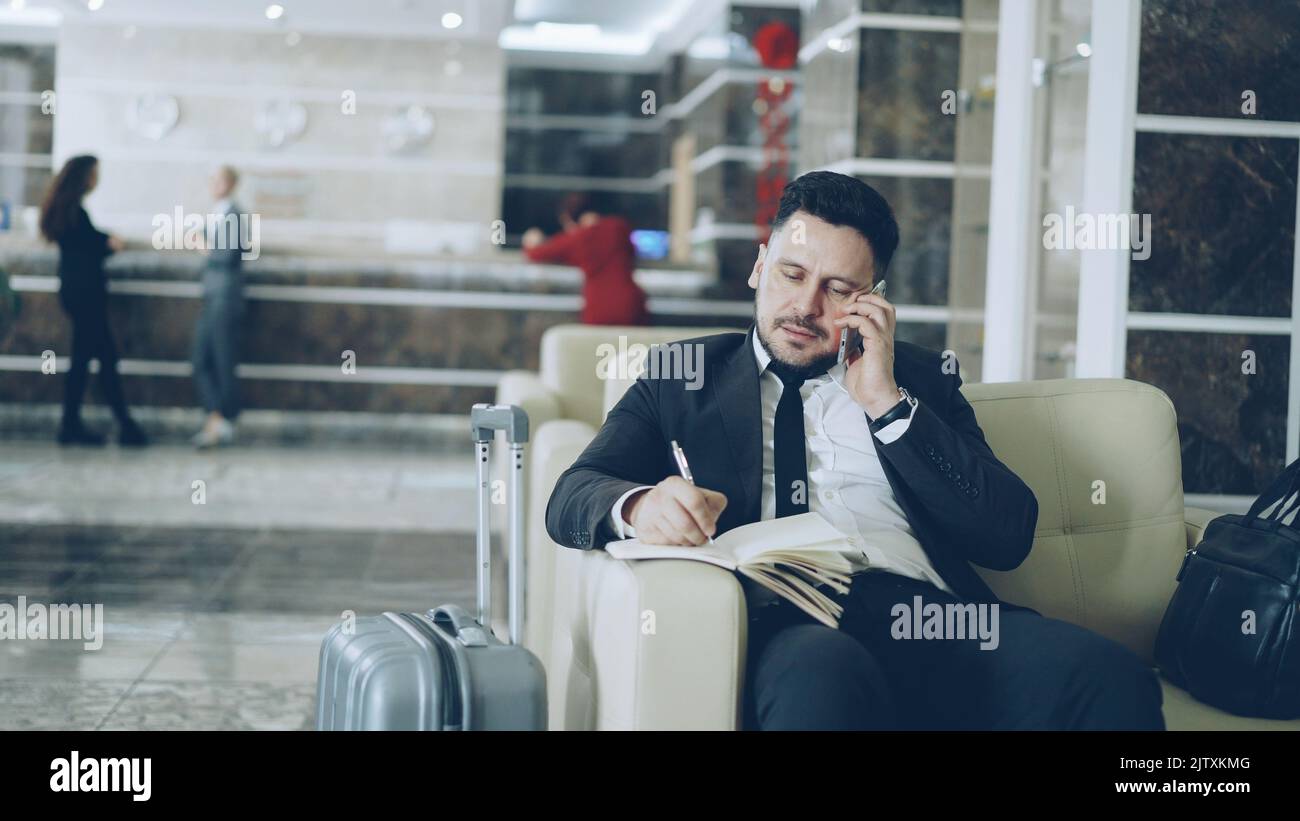 Pan shot di uomo d'affari fiducioso parlare telefono cellulare e scrivere note in blocco note mentre si siede sulla poltrona in hotel di lusso con bagagli vicino a lui. Concetto di viaggi, affari e persone Foto Stock