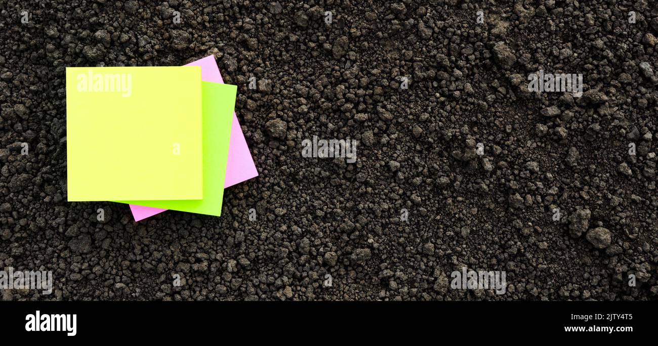 Giallo sticky note carta bianca terreno piano terra organica struttura. I colori notano il quadrato della carta sul terreno nero la struttura del terreno vicino in su. Mockup su suolo di terra Foto Stock