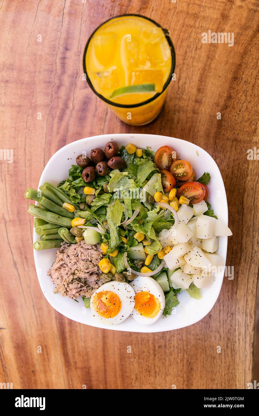 Insalata di tonno, uova, patate, lattuga, cipolla, pomodoro, mais, olive nere e asparagi. Servito in una ciotola bianca su un tavolo di legno. Bicchiere di succo d'arancia. Healt Foto Stock